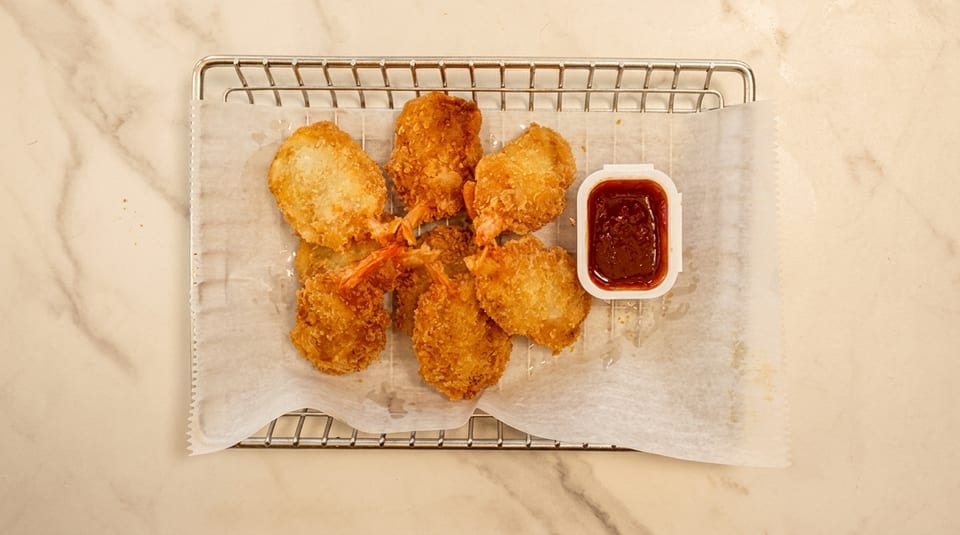 Golden brown fried shrimp with tails on, served on a metal rack with a side of dipping sauce.