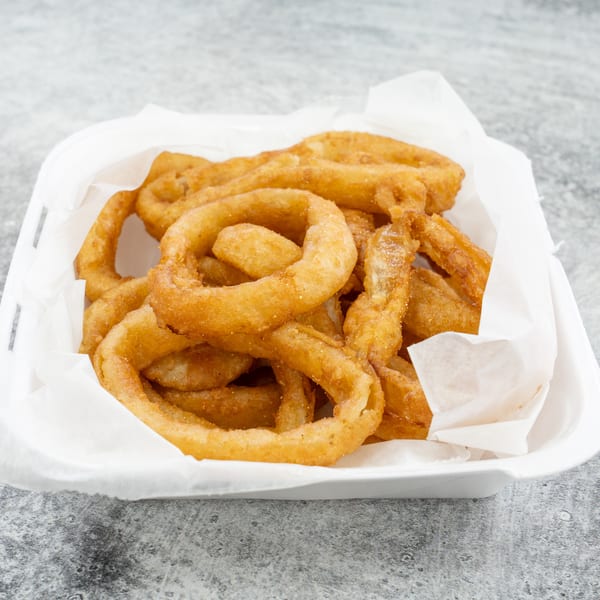 A white takeout container filled with golden-brown, crispy onion rings on a gray speckled surface.