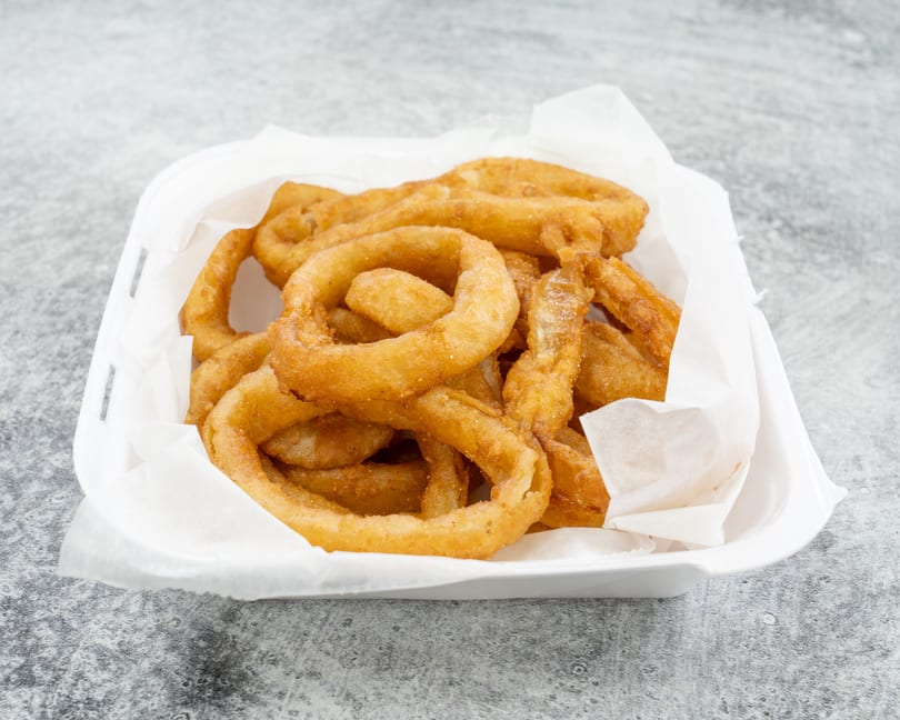 A white takeout container filled with golden-brown, crispy onion rings on a gray speckled surface.