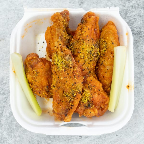 Top-down view of crispy orange chicken wings with yellow seasoning and celery sticks in a white takeout container.