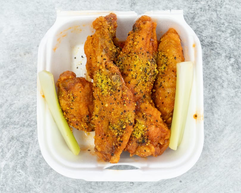 Top-down view of crispy orange chicken wings with yellow seasoning and celery sticks in a white takeout container.