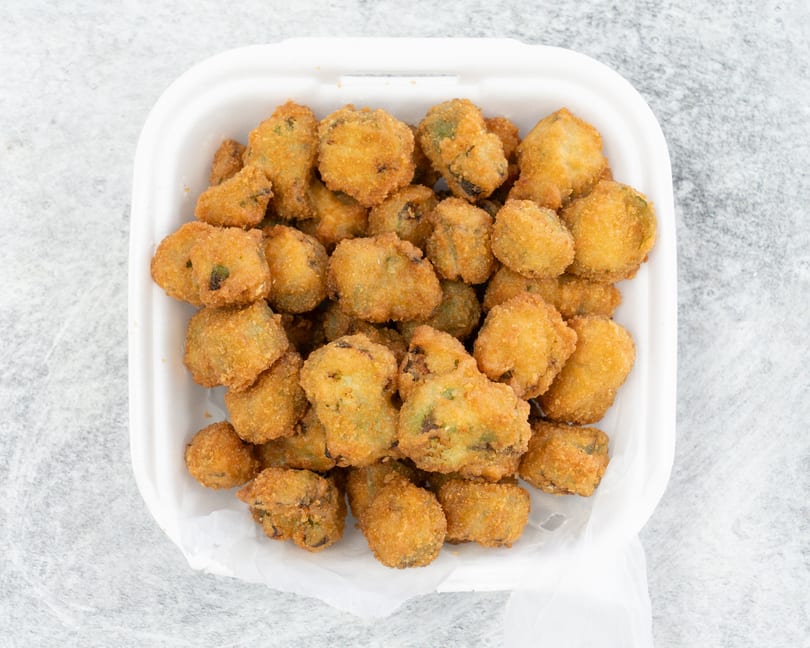 A white takeout container filled with golden-brown fried okra pieces on a light gray surface.