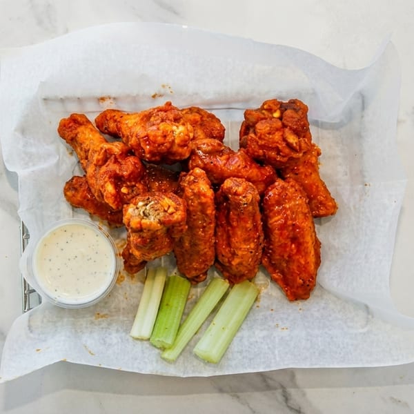 A basket of buffalo chicken wings with ranch dressing and celery sticks on a marble surface.